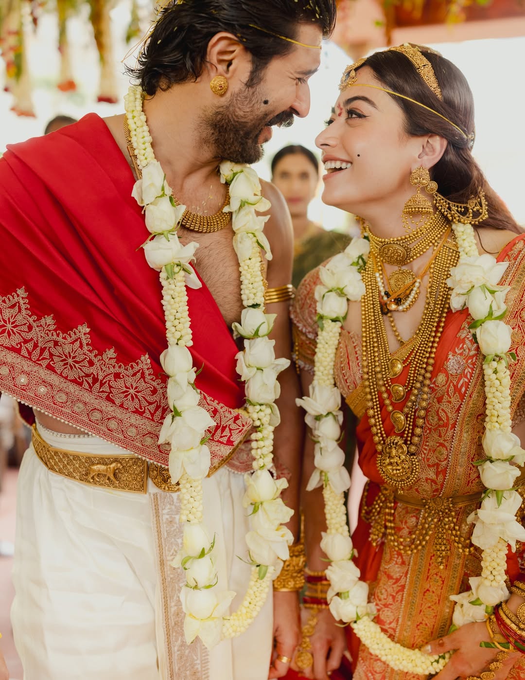 Vijay Deverakonda and Rashmika Mandanna smiling at each other wearing white rose garlands and traditional South Indian wedding attire